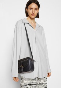 Camisa de corte holgado en gris claro con cuello, combinada con un bolso de hombro negro texturizado que tiene una solapa frontal y acentos en dorado.