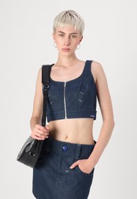 Denim krojeni top s sprednjim zadrgo, z modrim šivanjem in kvadratnim izrezom, v kombinaciji z denim mini krilom in črno torbico za čez ramo.