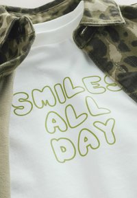 T-shirt blanc avec le texte « SMILES ALL DAY » en lettres bulles vertes, superposé sous une chemise en flanelle à imprimé léopard kaki et noir.