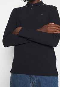 Man som bär en svart långärmad polotröja med en liten rosa logotyp på bröstet och blå jeans, med armarna över bröstet.