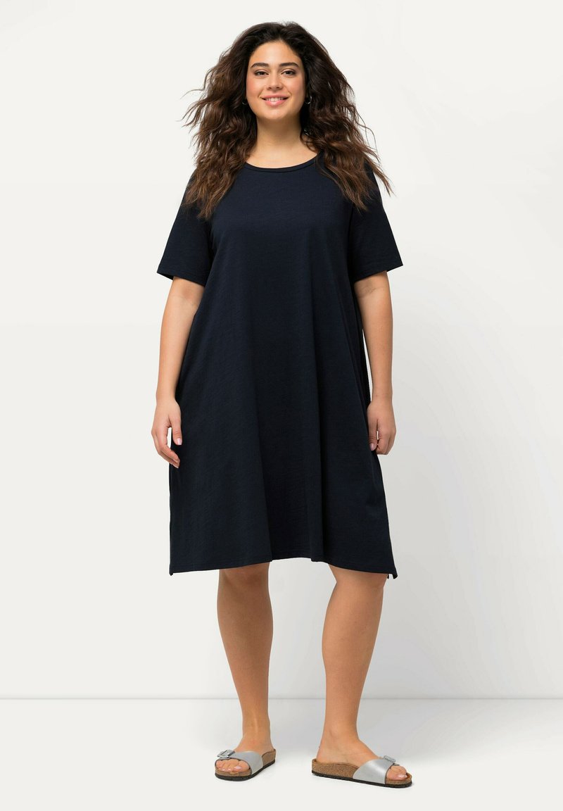 Nášivkově modré šaty s krátkým rukávem vyrobené z měkké tkaniny, s uvolněným A-liniovým střihem a bočními rozparky. Kombinované se šedými klouzavými sandály.
