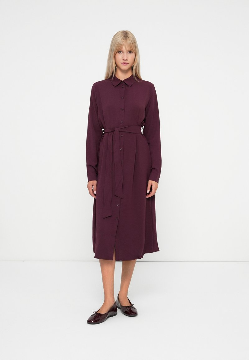 Burgunderfarget skjortekjole med knapper foran, lange ermer, midjebånd og sidespalter. Lett stoff med en glatt tekstur.