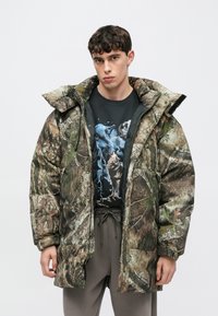 Giacca imbottita mimetica in tonalità di verde e marrone con collo alto, design oversize, chiusura con zip e grandi tasche.