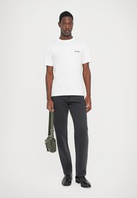 T-shirt bianco in cotone con piccola ricamo marrone sul petto, abbinato a jeans neri e scarpe nere, mentre tiene una borsa verde oliva.