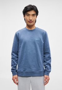 Sweatshirt azul com um tecido texturizado, decote arredondado, punhos ribados e um patch retangular da marca no peito.