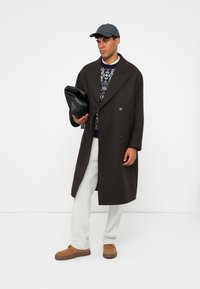 Uomo con un lungo cappotto marrone scuro, maglione a fantasia, pantaloni bianchi, scarpe beige e berretto nero, che tiene una pochette in pelle nera contro uno sfondo bianco.