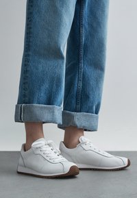 Persona, kas valkā zilas bikses ar aprocēm un baltas sporta kurpes ar brūnām zolēm, stāv uz pelēkas grīdas vienkāršas fona priekšā.