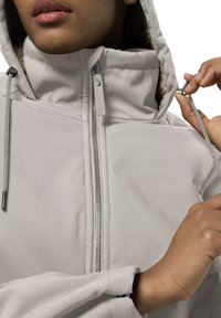 Osoba upravující šňůrku na kapuci světle šedé zipevé bundy s fleecovou podšívkou uvnitř kapuce.