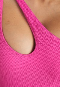 Lysende rosa badetøy med et teksturert, zigzag-mønster. Den har brede stropper og en dyp utringning, som fremhever et glatt og tettsittende design.