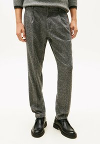 Pantalones grises, texturizados y de corte relajado, con plisados y cierre de botón en la cinturilla, combinados con zapatos negros.