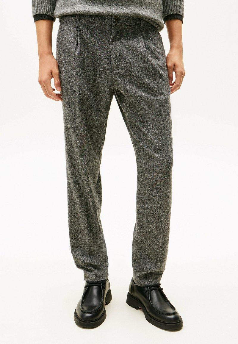 Pantalones grises, texturizados y de corte relajado, con plisados y cierre de botón en la cinturilla, combinados con zapatos negros.