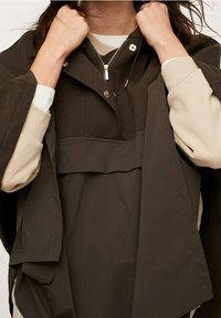 Vêtement d'extérieur marron surdimensionné avec un col montant, présentant une fermeture éclair avant et des boutons-pression. Fabriqué en tissu doux et texturé avec un design superposé.