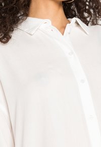 Vit, lätt skjorta med knappar som har en klassisk krage, subtil lyster och en avslappnad passform. Har klara plastknappar fram.