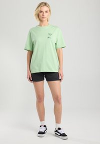 Light green cotton t-shirt with a relaxed fit, short sleeves, and a small graphic print. Paired with black denim shorts and sneakers.