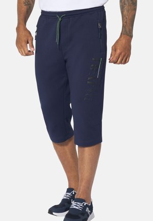 Mann trägt marineblaue knielange Shorts mit Reißverschlusstaschen und schwarz-weißen Turnschuhen, steht vor weißem Hintergrund.