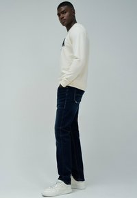 Sudadera crema con letras negras, jeans azul oscuro y zapatillas blancas. El modelo está de pie con las manos en los bolsillos frente a un fondo claro.