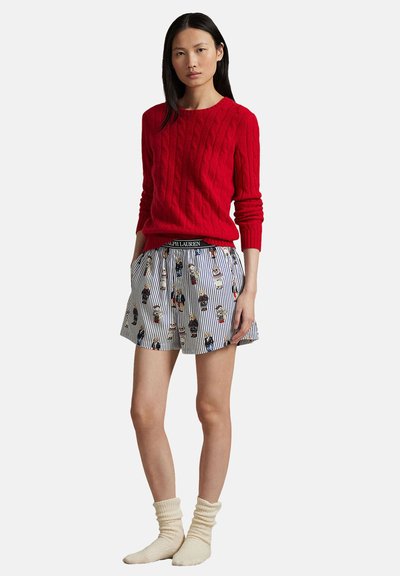 Röd kabelstickad tröja, randiga shorts med tecknad karaktärsprint och krämfärgade knähöga strumpor. Fokus på livfulla färger och texturkontrast.