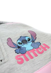 Sweat à capuche gris avec un personnage de dessin animé à la peau bleue et aux grandes oreilles, tenant en bas un texte rose "STITCH" en lettres grasses.