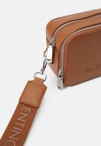 Borsa a tracolla in pelle marrone con doppie cerniere argentate, dettagli lucidi e una tracolla in tessuto con il nome del marchio stampato in marrone chiaro.