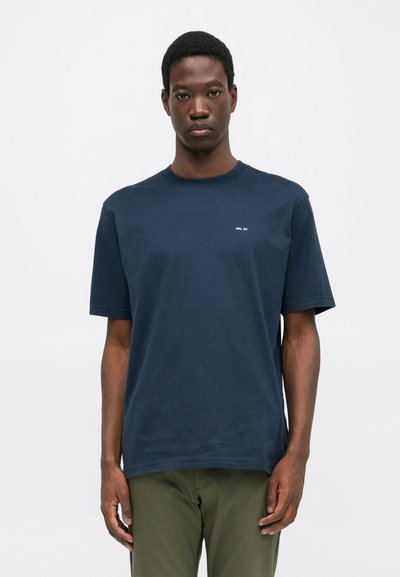 Junger Mann mit kurzen Haaren, der ein navyblaues T-Shirt mit kurzen Ärmeln und dem kleinen weißen Text "NN. 07" sowie olivgrünen Hosen vor einem weißen Hintergrund trägt.