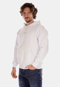 Felpa bianca con cappuccio, tasca frontale e logo testuale verde. Tessuto morbido, vestibilità comoda e cappuccio regolabile. Indossata sopra jeans blu.