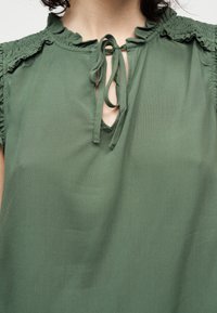 Blouse verte avec des épaules et un col à volants, ornée d'un nœud noué à l'avant avec une petite ouverture en forme de V.