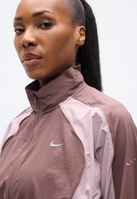 Ανεμοφράκτης Nike σε απόχρωση σκόνης τριανταφυλλιού και ανοιχτού ροζ, με ψηλό γιακά, κλείσιμο με φερμουάρ και λογότυπο στο στήθος. Υφή υφάσματος.