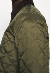 Olivengrön vadderad jacka i satinmaterial, med en mörkbrun cordflorskrage och ett diamantformat sömnadsmönster.