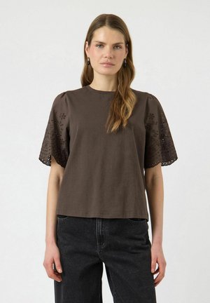 Vrouw met lang lichtbruin haar draagt een donkerbruin kortärmelig shirt met bloemvormige uitsnijdingen en zwarte high-waisted jeans, staat neutraal.