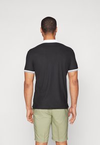 Melnā poloshirt ar balto apkakli un piedurkņu akcentiem, izgatavots no teksturētas auduma. Valkāts ar gaiši zaļām šortiem; redzama aizmugure.