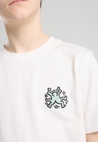 T-shirt bianco in cotone con un piccolo ragno cartoon ricamato in nero e verde menta sul lato sinistro del petto. Maniche corte e collo rotondo.