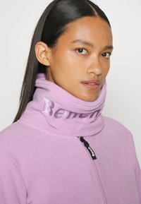 Lila Fleecejacke mit hohem Kragen und einem glänzenden Logo. Der Reißverschluss und die weiche Textur sorgen für Wärme und Komfort.