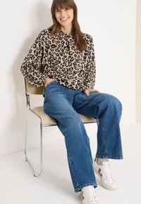 Langärmlige Bluse mit Leopardenmuster, hellbraun mit schwarzen Flecken, kombiniert mit blauen Jeans mit weitem Bein und weißen Turnschuhen mit roten Akzenten.