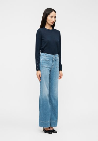 Μπλε ναυτικό πουκάμισο με μακρύ μανίκι και διακοσμητικά κουμπιά, συνδυασμένο με ανοιχτό μπλε τζιν παντελόνι με φαρδύ πόδι, ψηλό ύψος μέσης και πίσω τσέπες, φορεμένο με μαύρα μυτερά παπούτσια.