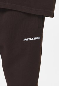 Pantalones de chándal marrones con una textura suave que incluyen un logo blanco de "PEGADOR" impreso en el muslo izquierdo. Incluyen bolsillos laterales para almacenamiento.