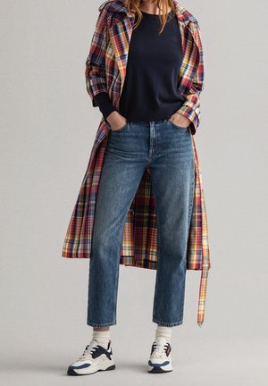 Personne portant un manteau à carreaux coloré par-dessus un pull noir, un jean bleu, des chaussettes blanches et des baskets blanches avec des accents bleu marine et rouge.