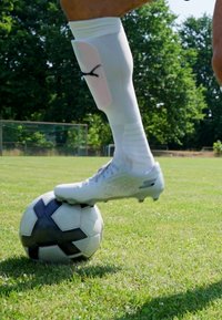 Crampons de football blancs avec une surface texturée posés sur un ballon de football noir et blanc sur de l'herbe verte, avec des arbres et un poteau de but en arrière-plan.