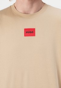 Beige tričko s kulatým výstřihem a krátkými rukávy. Obsahuje červenou obdélníkovou nášivku s nápisem "HUGO" v černém písmu. Materiál ze směsi bavlny.