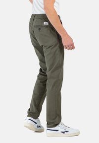 Pantalones de algodón verde oliva con cintura elástica y un bolsillo trasero, que incluye una pequeña etiqueta. Combinados con zapatillas blancas.