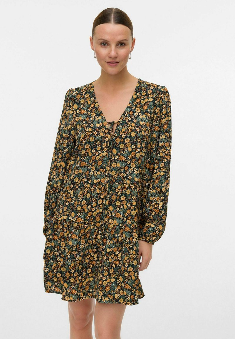 Femme portant une robe à manches longues, imprimée florale, avec un décolleté en V et un détail à nouer, se tenant contre un fond clair et uni.