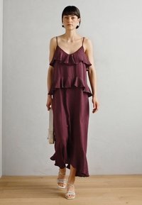 Bordeauxfarbenes ärmelloses Kleid mit geschichteten Rüschen am Oberteil, fließendem Rock und leicht gewelltem Saum. Kombiniert mit weißen Sandalen mit Riemchen.