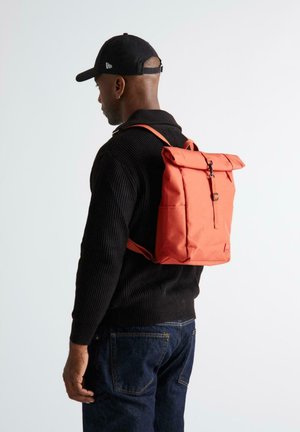 Hombre con suéter negro, gorra negra y jeans oscuros, con una mochila de rollo naranja brillante, de perfil sobre fondo sencillo.