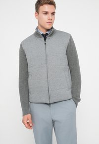 Γκρι γιλέκο με φερμουάρ και επενδεδυμένο μπροστινό μέρος, συνδυασμένο με πλεκτά μανίκια. Διαθέτει όρθιο γιακά και πλαϊνές τσέπες, συνδυασμένο με ανοιχτό γκρίζες παντελόνες.