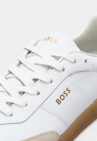 Bílé kožené tenisky s béžovými semišovými detaily, bílými šňůrkami a zlatým logem "BOSS" po boku a na jazyku, s hnědou podrážkou.