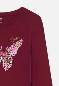 Bordo pitkähihainen paita, jossa on kukallinen perhosdesign vaaleanpunaisena, vihreänä, sinisenä ja kultahippuina, sekä sana "Rakkaus" kultaisella käsinkirjoituksella sen yläpuolella.