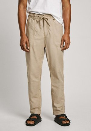 Beige cotton blend pants with an elastic waistband and drawstring. Features two side pockets and tapered legs. Worn with black sandals.
