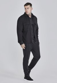 Chaqueta de camisa negra con dos bolsillos, camisa negra de manga larga, pantalones oscuros y zapatillas negras. Textura suave y detalles de diseño elegantes.