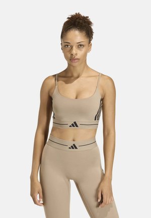 Mujer con pelo rizado lleva un sujetador deportivo beige y leggings de cintura alta con el logo negro de Adidas y rayas sobre un fondo blanco.