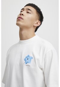 Hvid bomulds t-shirt med et lysblåt blomsterdesign på venstre bryst. Korte ærmer, rund halsudskæring, afslappet pasform.