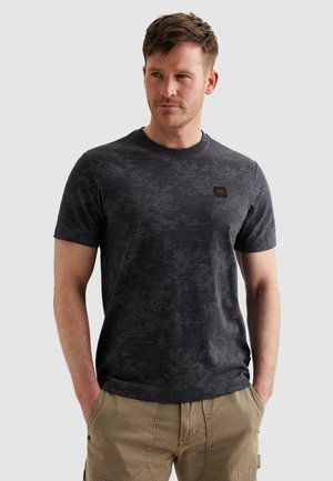 Muž s krátkymi vlasmi, oblečený v tmavosivej tričku so vzorom a béžových cargo šortkách, stojaci s rukami v vreckách a pozerajúci sa doprava.